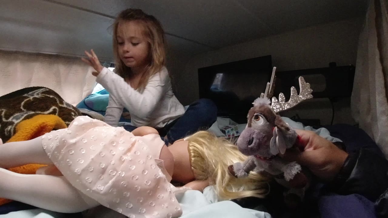 In Santa Maria, California, the young girl raises one hand while sitting cross-legged on the bed, with the doll and plush reindeer toy still in the foreground.