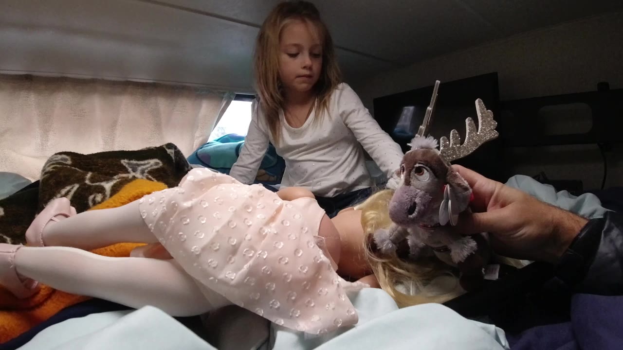 At 0:00 in Santa Maria, California, a young girl sits on a bed inside a vehicle while an adult hand holds a stuffed reindeer beside a doll in a pink dress.