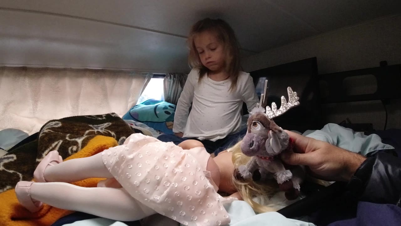 In Santa Maria California, a young girl sits on bedding behind a doll in a pink dress as an adult hand brings a stuffed moose toward the doll's face.