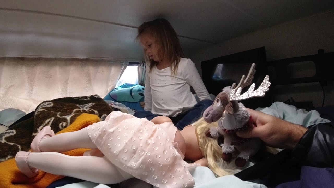 In Santa Maria California, the girl turns toward the right side of the bed as the stuffed moose remains near the doll lying across the bedding.