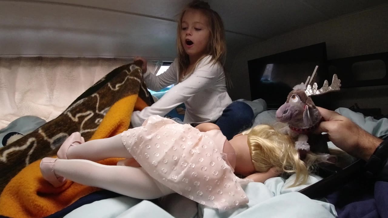 In Santa Maria California, a young girl sits on a bed inside a vehicle interior while a blonde doll lies across blankets in the foreground and a plush toy is held near her.