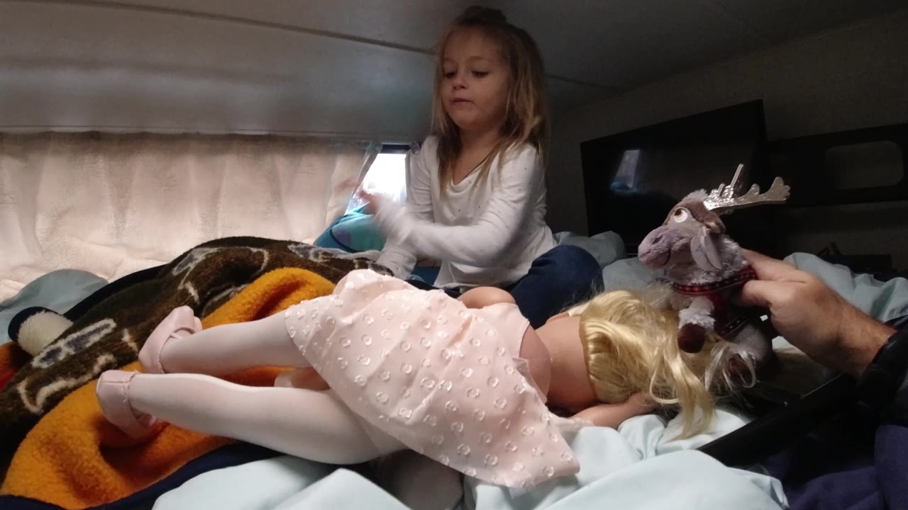 In the vehicle sleeping area, the child sits upright behind the doll while the plush toy shifts to the right side of the frame near the doll's head.