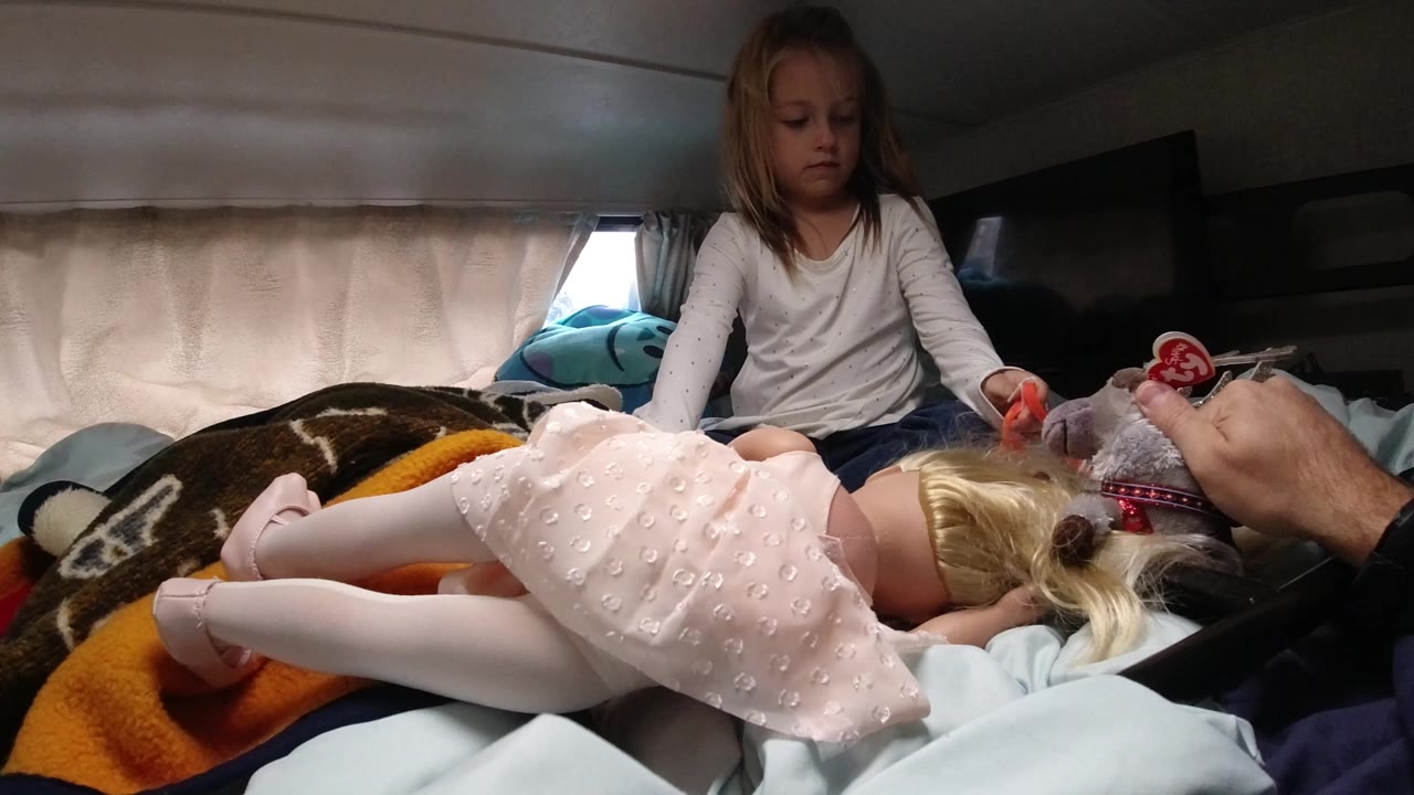 In Santa Maria, California, a young girl sits on a bed inside a compact vehicle-like interior while a blonde doll in a pink dress lies in the foreground and an adult hand holds a small toy near the doll.