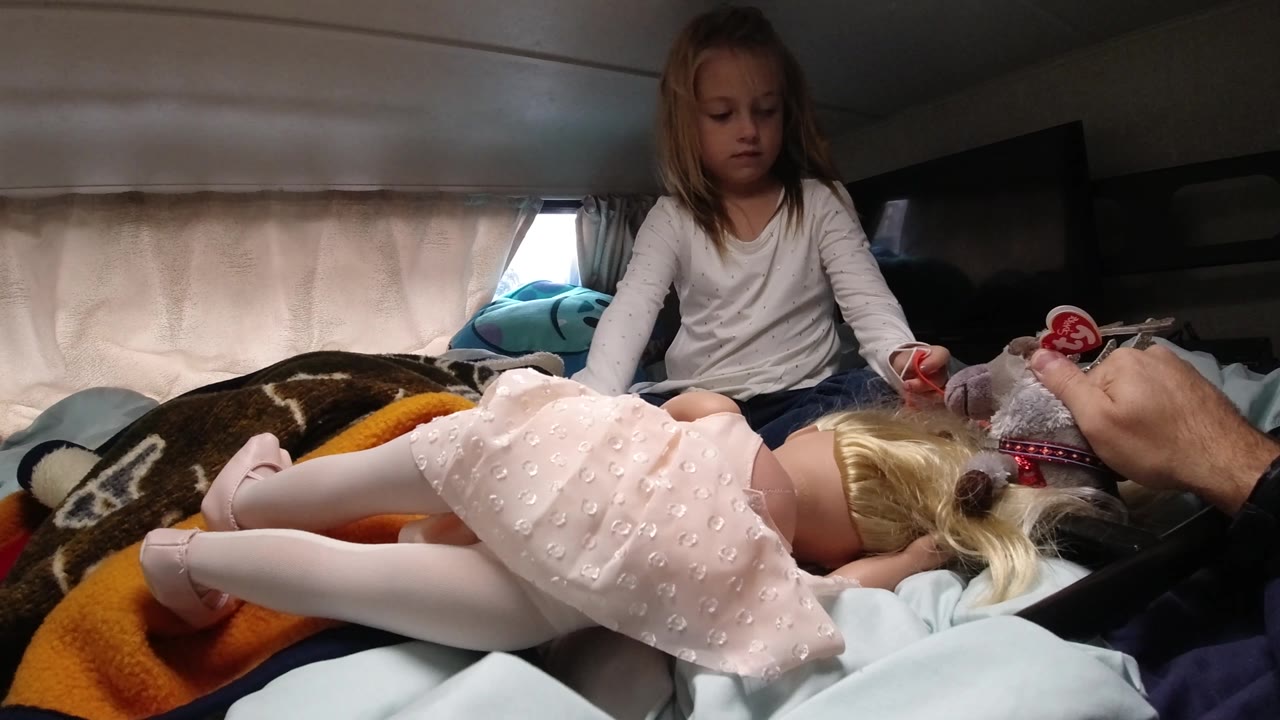 A young girl sits upright behind the doll, with blankets and a curtained window around the bed and an adult hand holding a small toy close to the doll in the foreground.