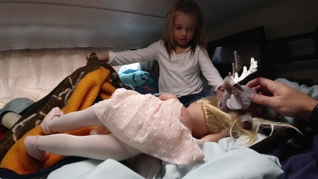 In Santa Maria, California, a young girl sits on a bed while a blonde doll lies across blankets and an adult hand holds a stuffed moose near the foreground.