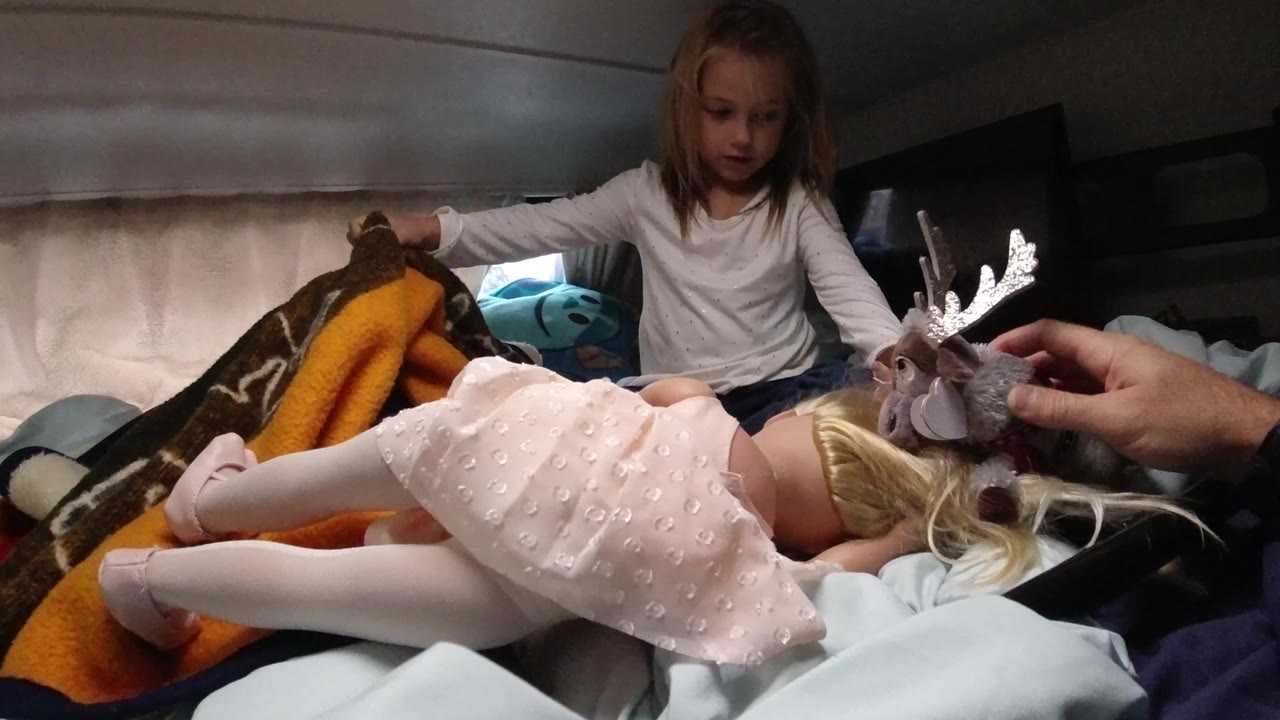Girl watches as an adult hand holds a stuffed moose beside a doll on the bed.