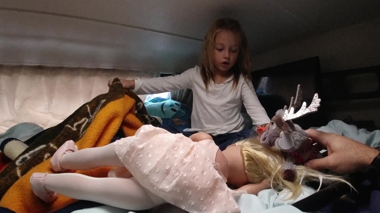 In Santa Maria, California, the girl remains seated behind the doll while the moose toy is presented near the doll’s hair and the pale curtains glow softly behind her.