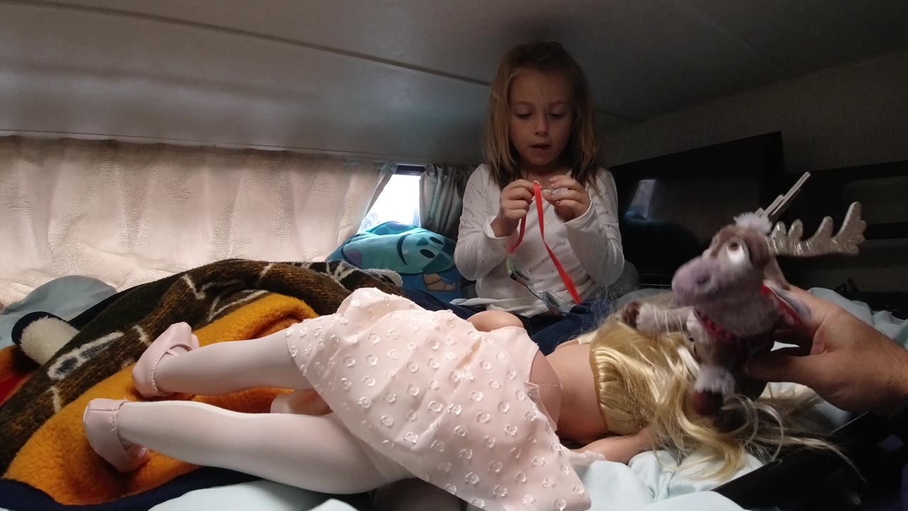 In Santa Maria, California, the girl continues handling the red strip while the stuffed reindeer faces toward the blonde doll lying across the lower frame.