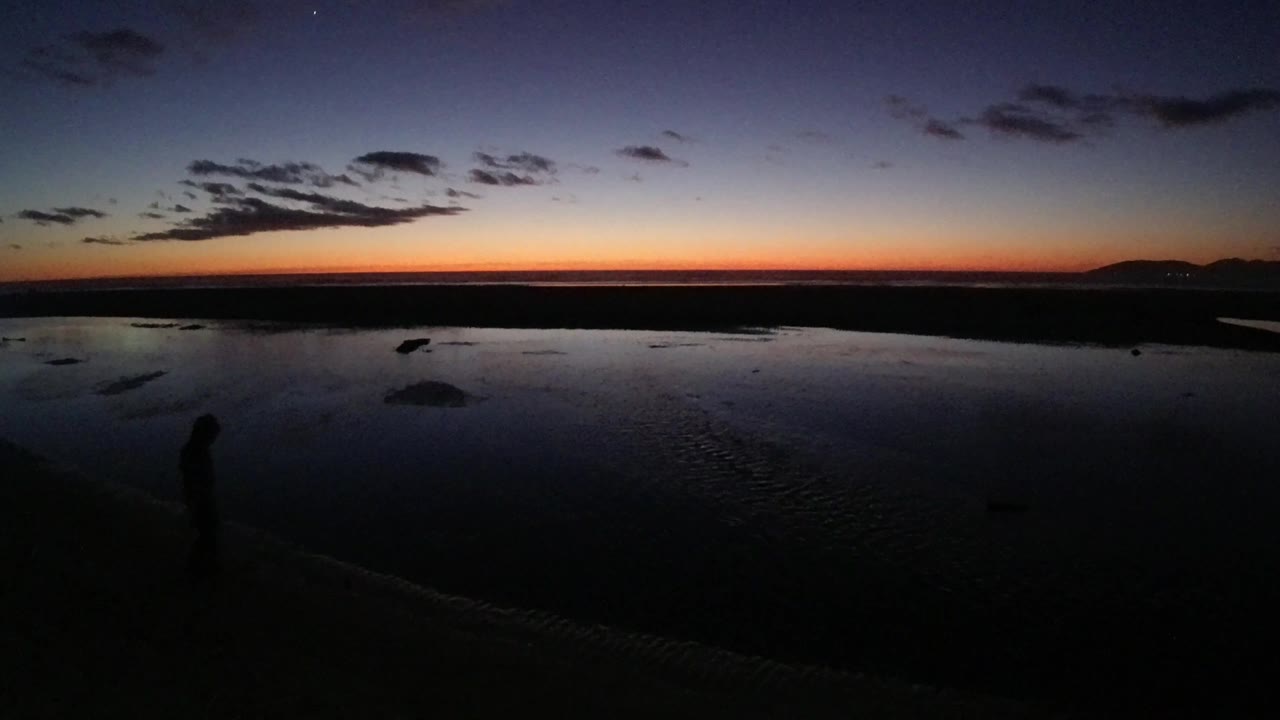 Dark beach foreground, reflective water, orange horizon, and a small standing silhouette.