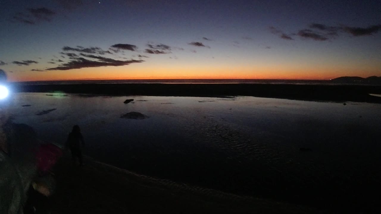 Pismo Beach California twilight frame with a bright light flare near a person at the left side, reflective water, dark sand, distant land, and an orange horizon under blue sky.
