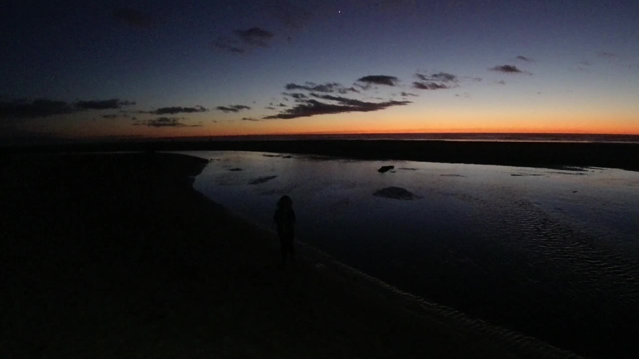 Wide twilight view at Pismo Beach with a silhouetted woman low in frame, dark sand in the foreground, reflective water beyond her, and an orange band along the horizon.