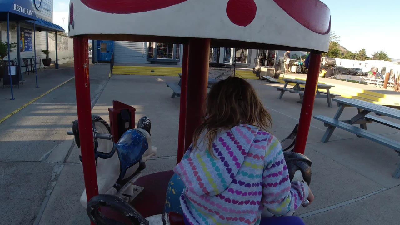 A young blonde girl in a colorful striped hoodie is seen from behind on a carousel horse in Pismo Beach, California, with red poles, picnic tables, and storefronts ahead.