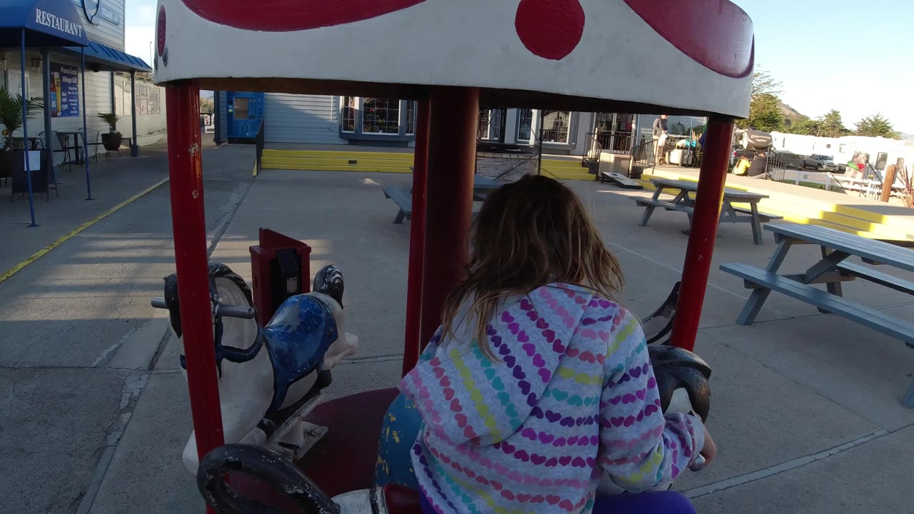 Girl rides beneath red carousel poles with picnic tables and storefronts ahead.