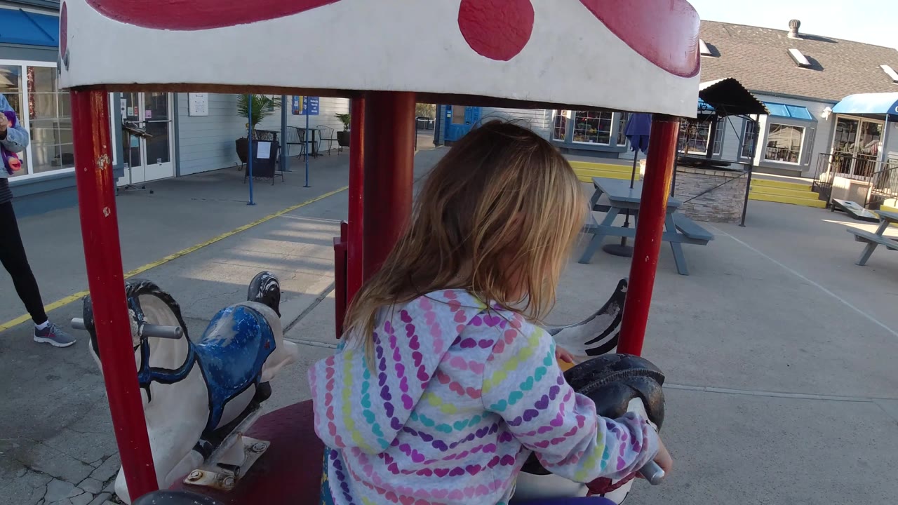The young girl is shown in side profile on the carousel in Pismo Beach, California, with her striped hoodie, blonde hair, red poles, and nearby storefront windows visible.