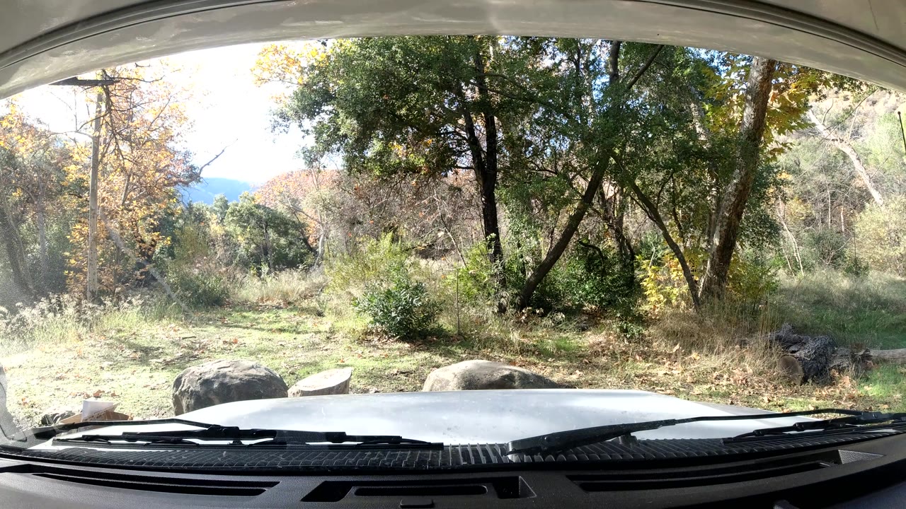 At 0:02, sunlight brightens the right side of the Ojai California woodland view while the car hood, wipers, rocks, grass, and trees remain in frame with no visible people.