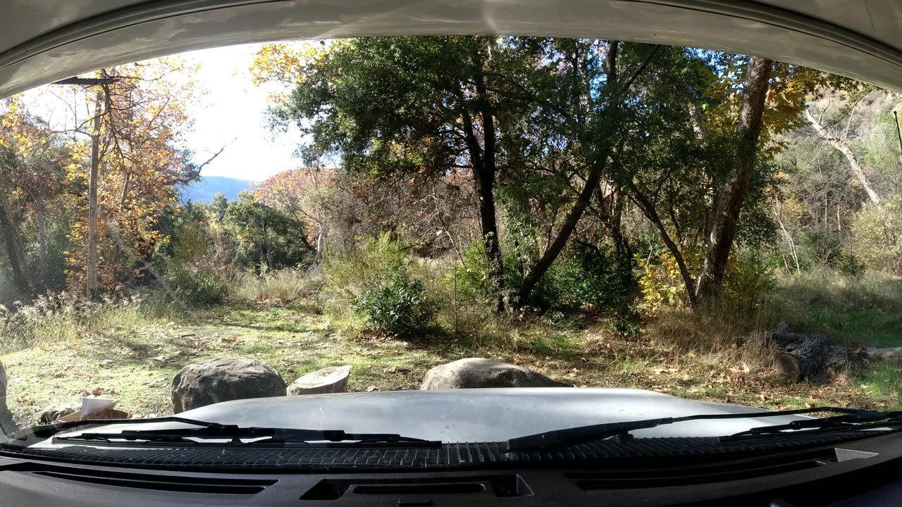 At 0:03, the static car POV continues with the hood and wipers along the lower edge, large rocks near the clearing, layered trees behind them, and no visible people.