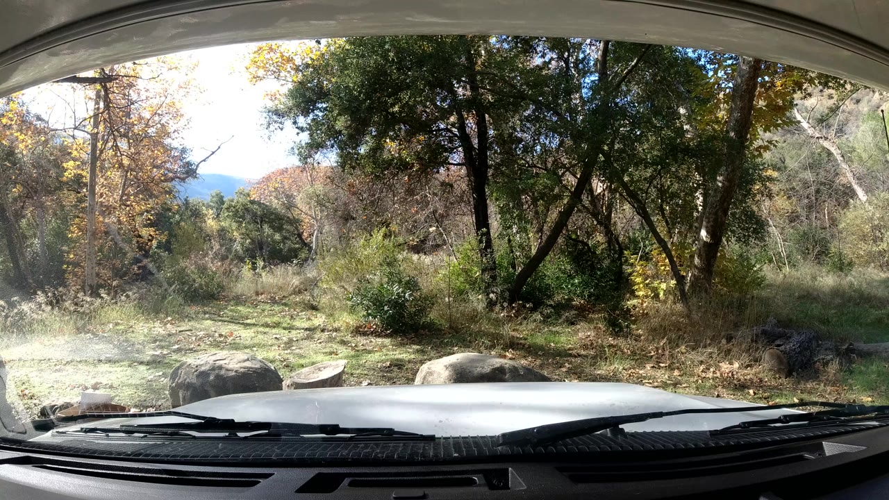 Opening frame from inside a vehicle in Ojai California, looking past the hood and wipers toward sunlit trees, dry grass, shrubs, and rocks with no visible people.