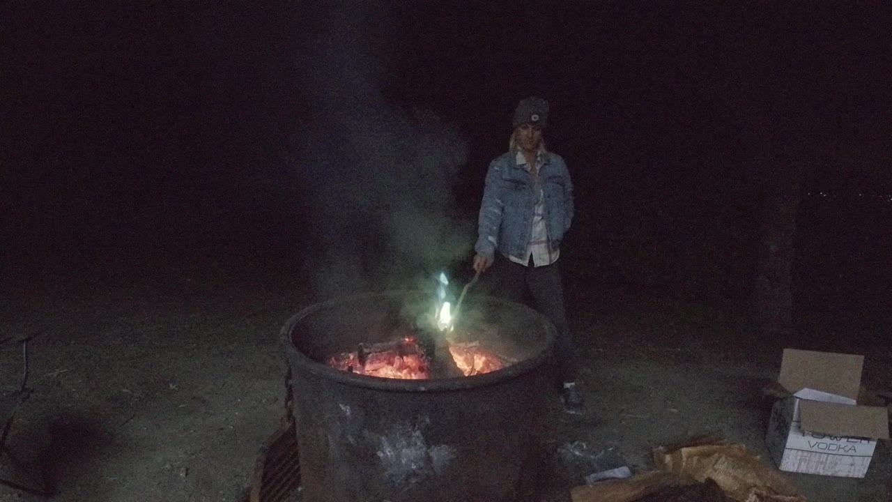 A man in a denim jacket and cap stands behind a large metal fire pit in San Clemente California at night while embers glow and smoke rises.