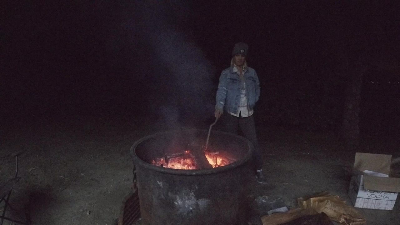 A man stands upright behind the smoky fire pit at night, with a cardboard box on the ground at frame right and red embers visible inside the metal ring.