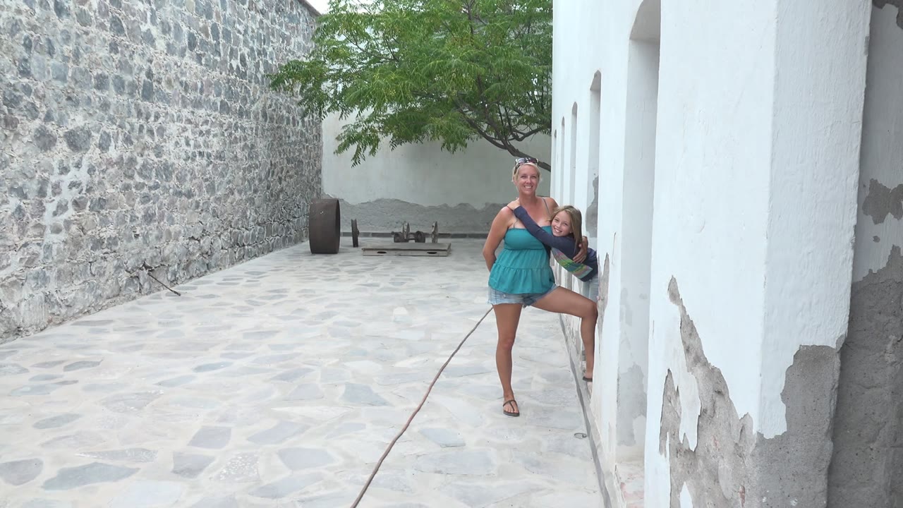 At the start of the Mulege BCS Mexico clip, a woman in a blue dress holds a child near a white building in a pale stone courtyard.