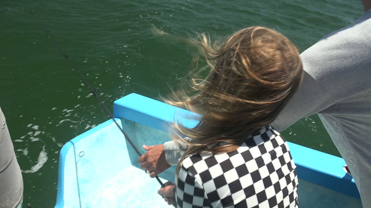 At 0:00 in La Bocana, BCS, Mexico, a young girl with windblown hair faces away beside the bright blue boat edge while an adult arm and fishing rod appear over green water.
