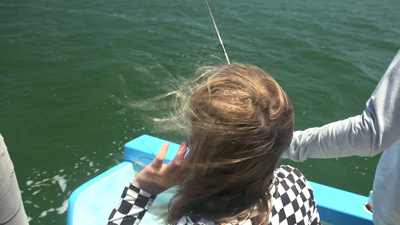 At 0:02 in La Bocana, BCS, Mexico, the girl faces down toward the water beside the blue boat edge while a partially seen adult in a gray shirt stands at the right.