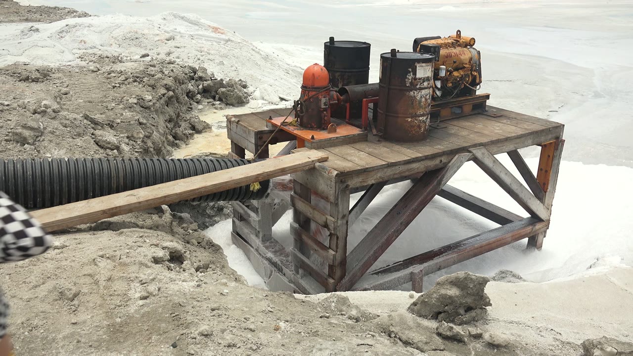 A blurred checkered sleeve enters the foreground at the edge of the Guerrero Negro BCS Mexico pump setup, with the wooden platform, barrels, pipe, and muddy water still visible.