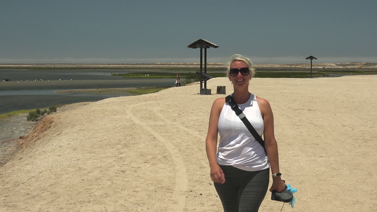 At 0:00 in Guerrero Negro BCS Mexico, a woman in sunglasses walks toward the camera on a pale sandy path with water to the left and small roofed pavilions in the distance.