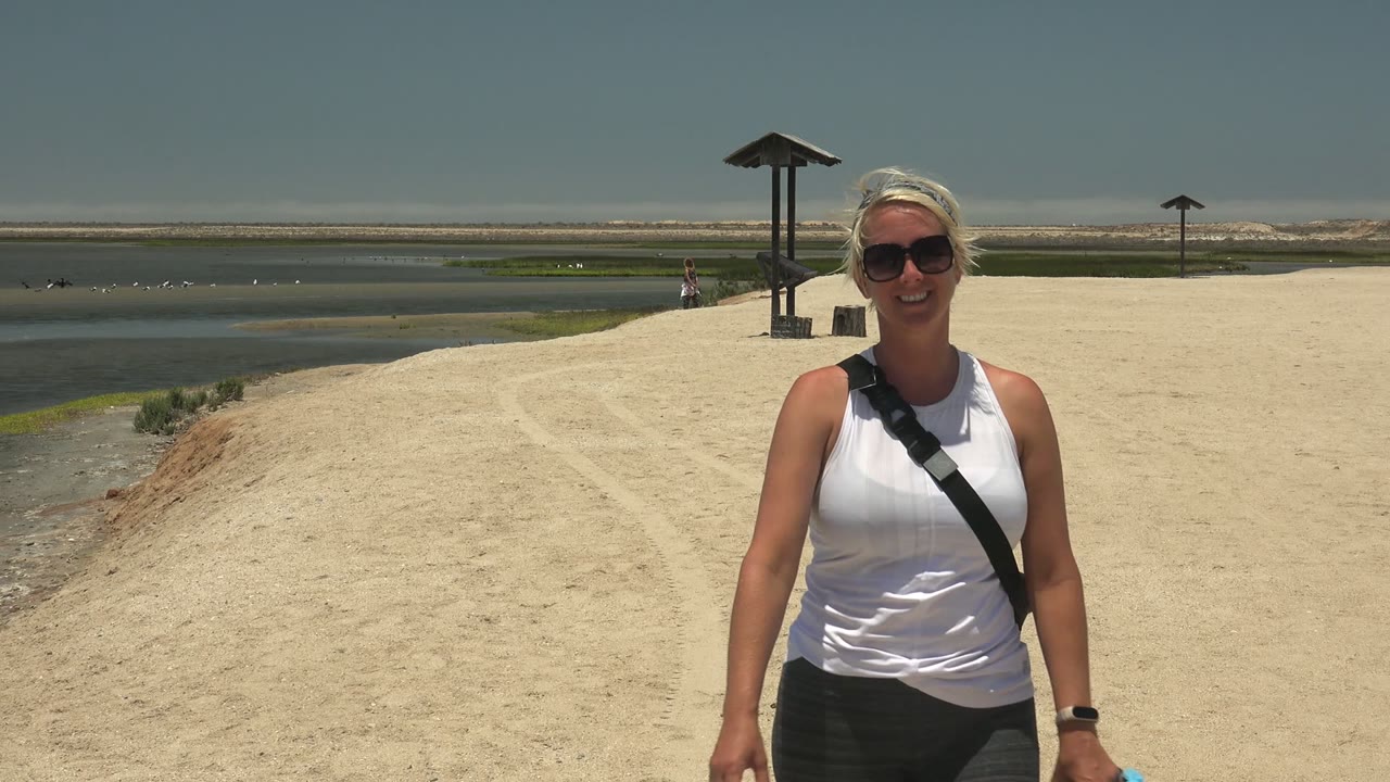 Woman closer to camera on sandy path with water and pavilions behind.