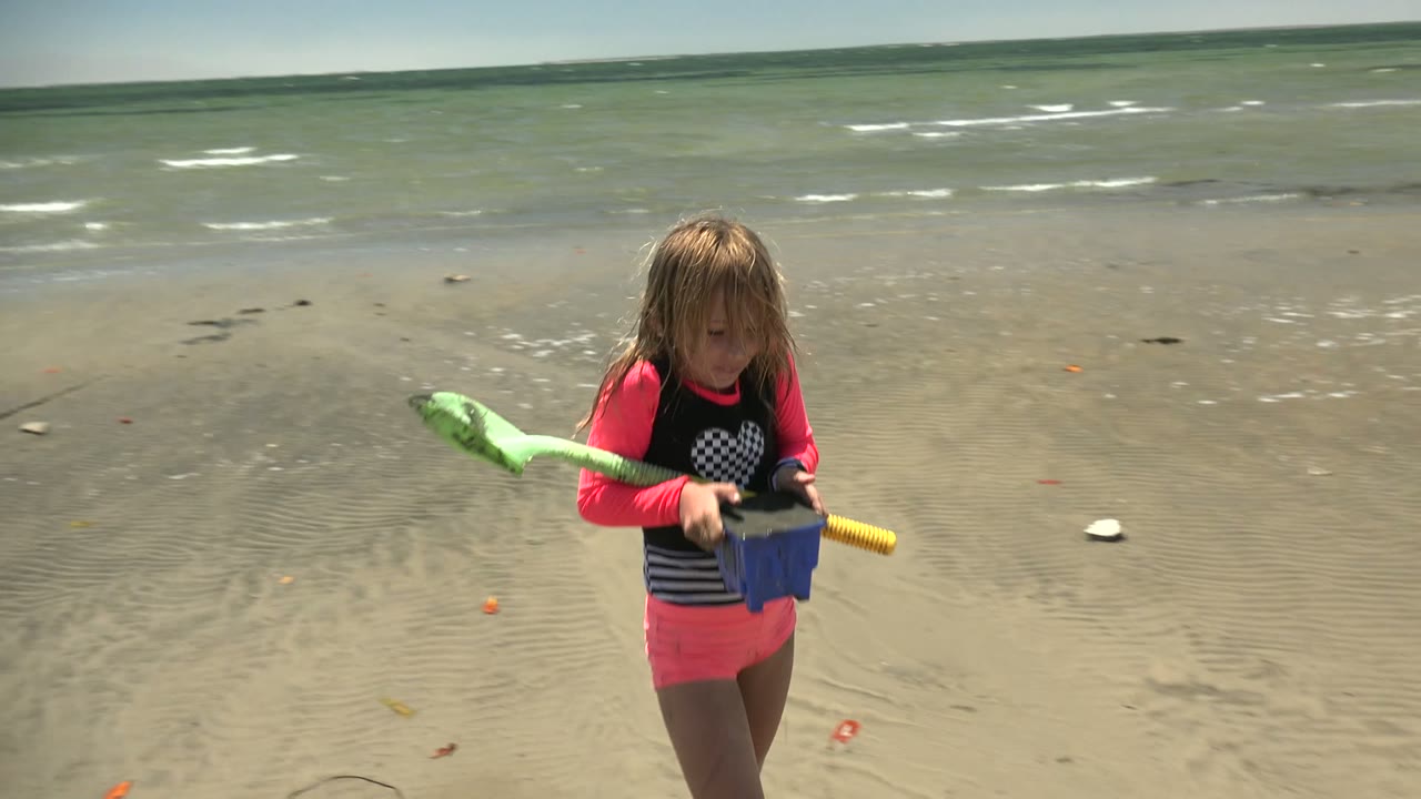 A child in a bright pink and black swim shirt stands on wet sand in San Carlos BCS Mexico holding a green shovel and blue bucket, with shallow ocean waves behind them.