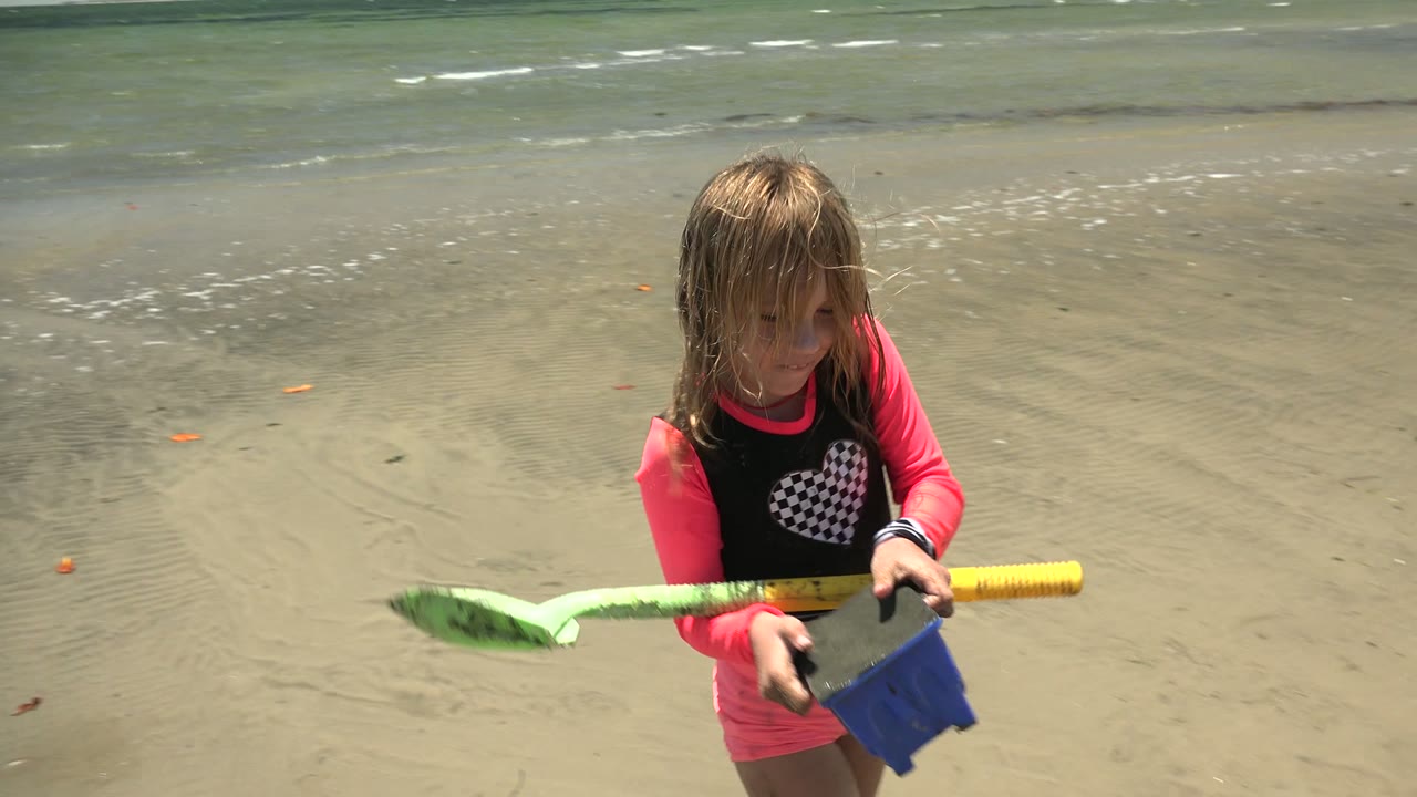 Child looking down while holding a shovel and bucket on wet sand.