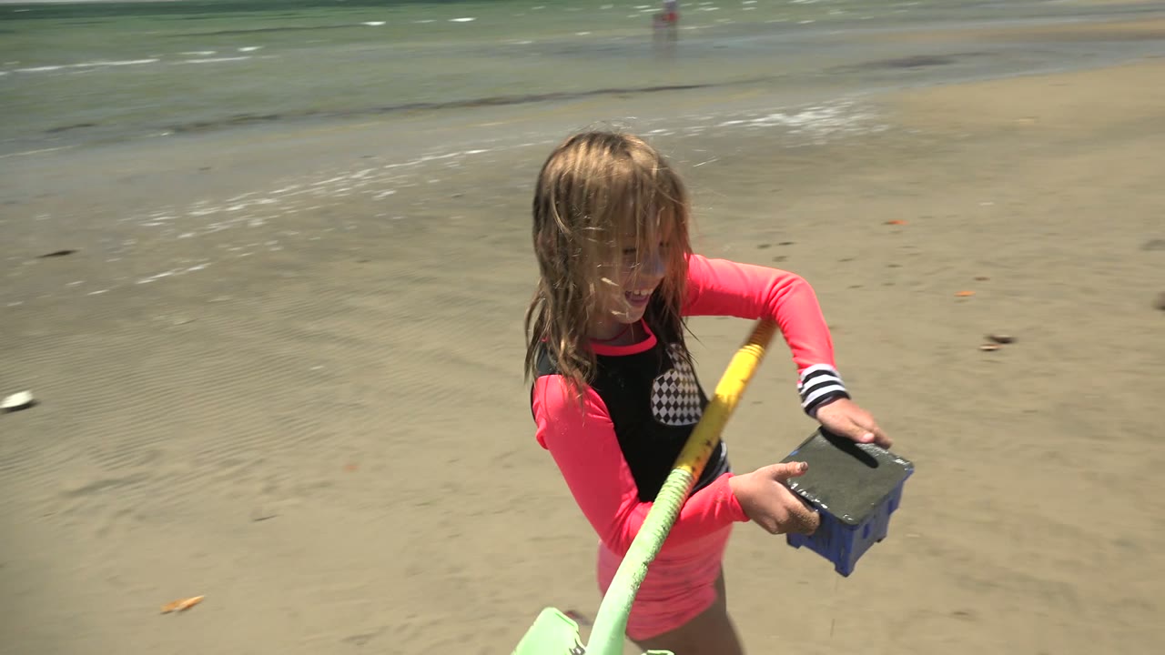 The child turns slightly to one side on the sandy San Carlos BCS Mexico shore, holding the blue bucket and green shovel while the low surf runs across the background.
