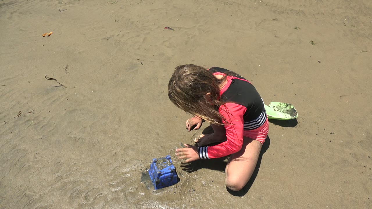 At 0:00 in San Carlos BCS Mexico, a child kneels on wet beach sand with hands near a blue sandcastle mold while a green sand toy sits behind them.