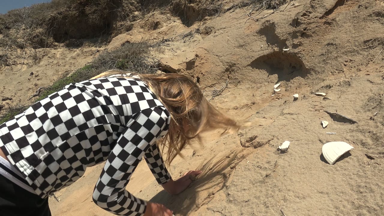 At 0:02 in San Quintin BC Mexico, the person leans low over the ground, with hair falling forward and hands close to small pale fragments embedded in the sand.
