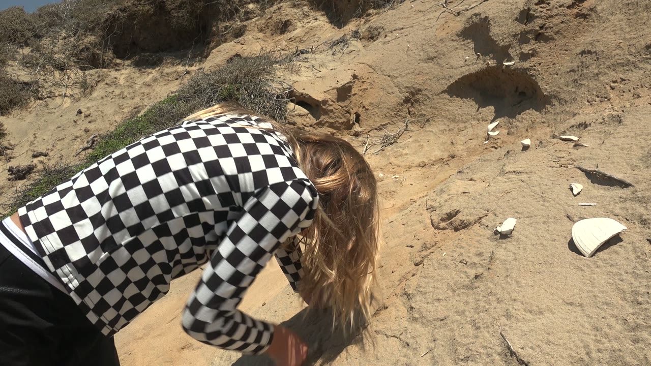 At 0:02 in San Quintin, Mexico, the subject's back and checkered shirt dominate the foreground while sandy ground and sparse scrub fill the background.