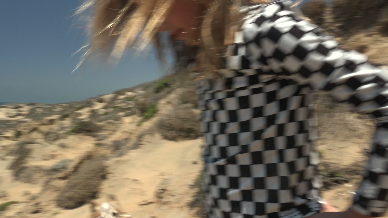 At the start of the San Quintin, Baja California, Mexico clip, a close blurred woman in a checkered or striped top passes near the camera with sandy bluffs and ocean behind her.