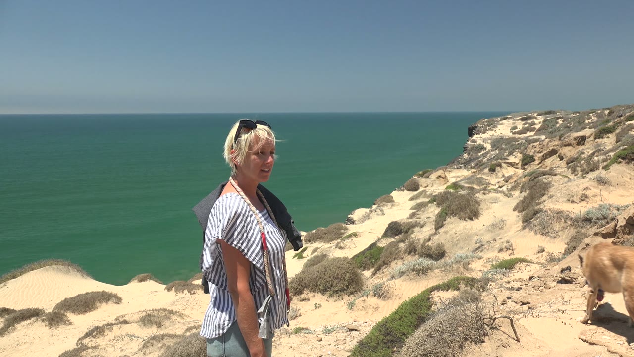 Blonde woman in a striped top stands on a sandy coastal cliff path in San Quintin, Mexico, with green ocean behind her and a dog partly visible at the right edge.