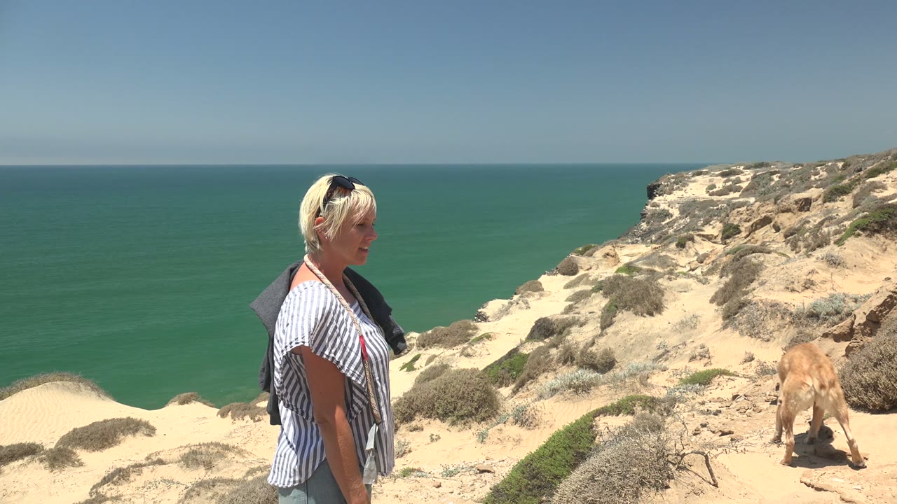 Woman walking along sandy coastal path as dog moves ahead on right.