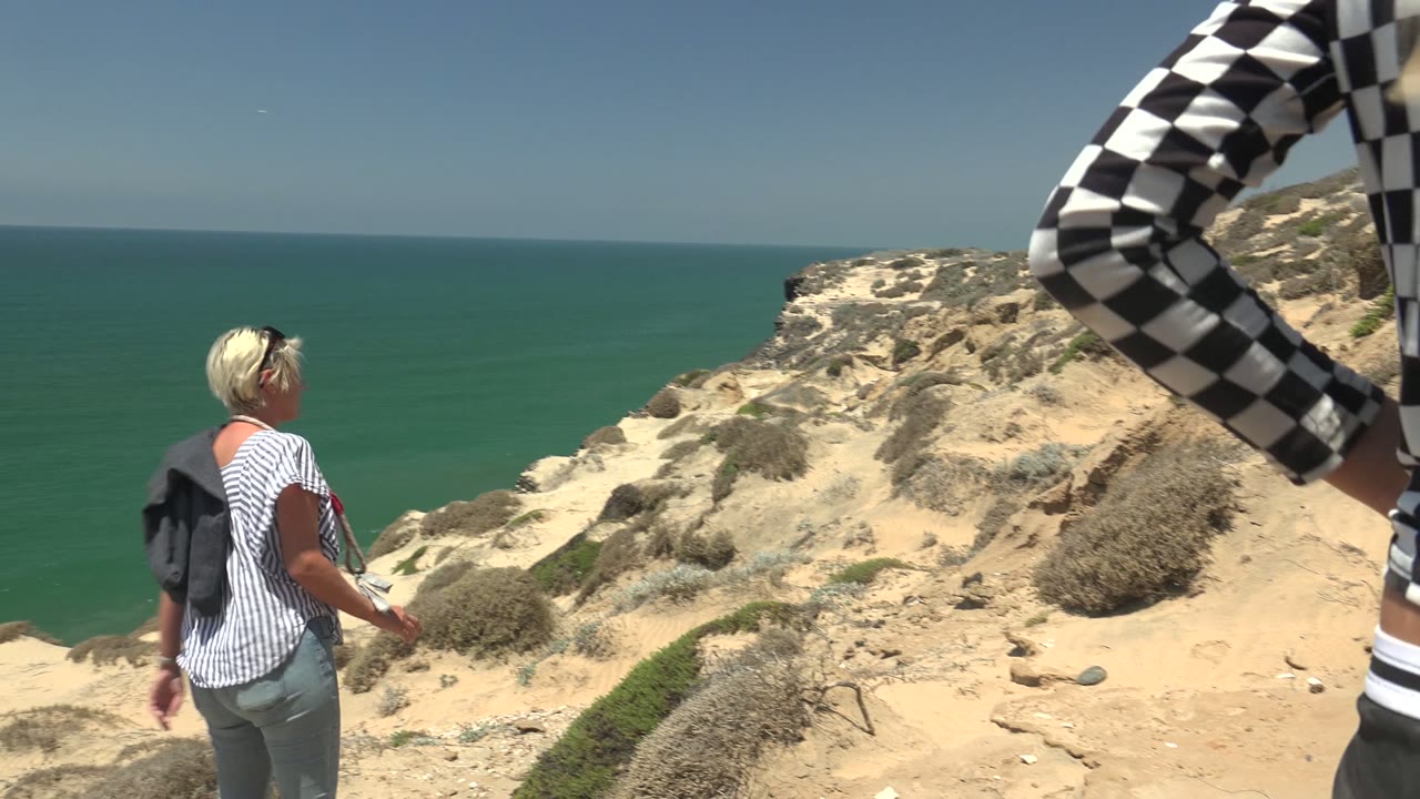 A woman stands near the edge of a sandy coastal cliff in San Quintin, Mexico, looking toward the ocean while a cropped person in a checkered shirt enters the right edge of the frame.