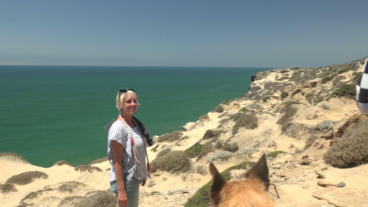 A woman stands on the sandy San Quintin cliff with the dog's head partly visible at the bottom of frame, backed by scrub-covered rock and green-blue ocean.