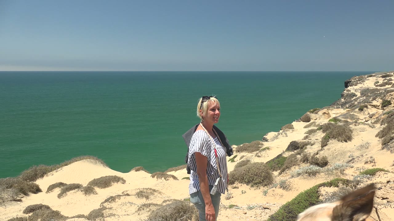 A blonde woman stands on a sandy coastal cliff in San Quintin BC Mexico with green-blue ocean and clear sky behind her.