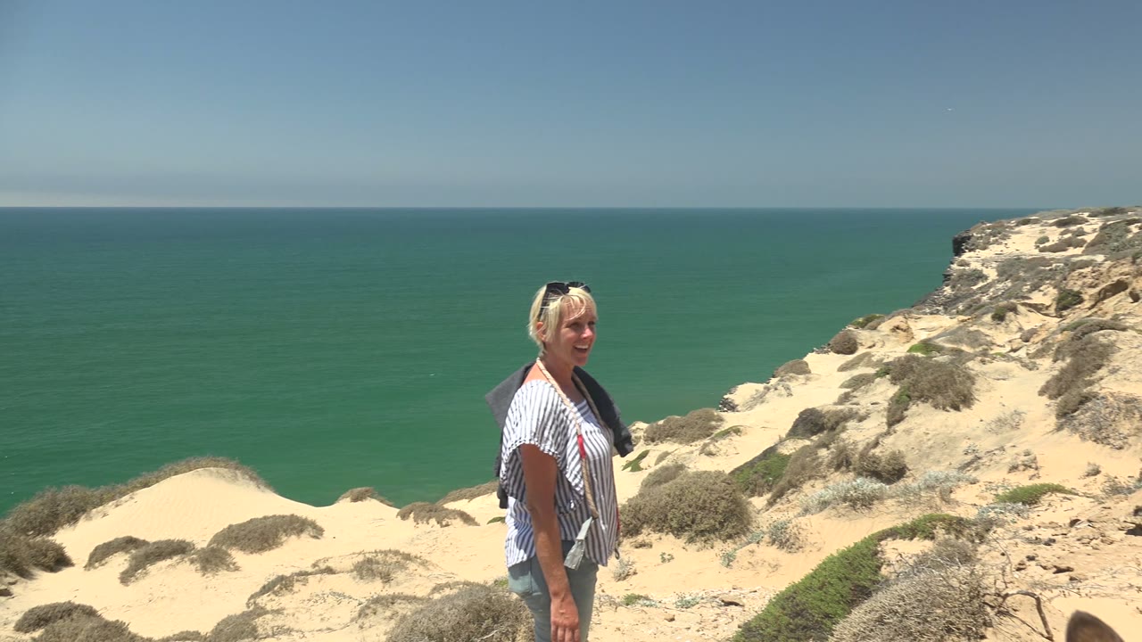 The woman remains in profile near the sandy bluff in San Quintin BC Mexico, with a broad ocean horizon and scrub-covered cliff behind her.