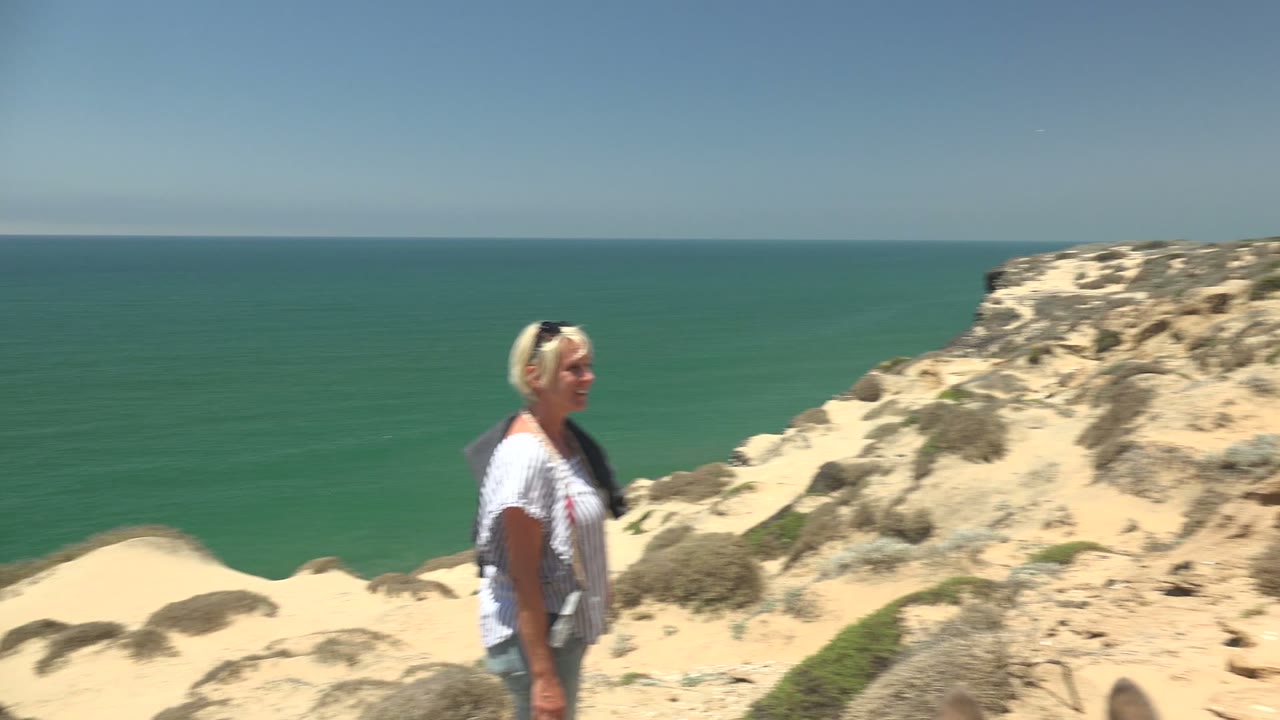 The woman is slightly blurred as she moves along the sandy San Quintin cliff, with ocean water on the left and pale dunes on the right.