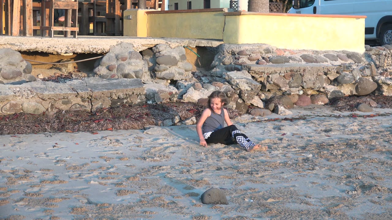 At 0:00, a child sits low on the sandy beach in San Carlos BCS Mexico with rocks, seaweed, a low wall, and wooden furniture behind her.