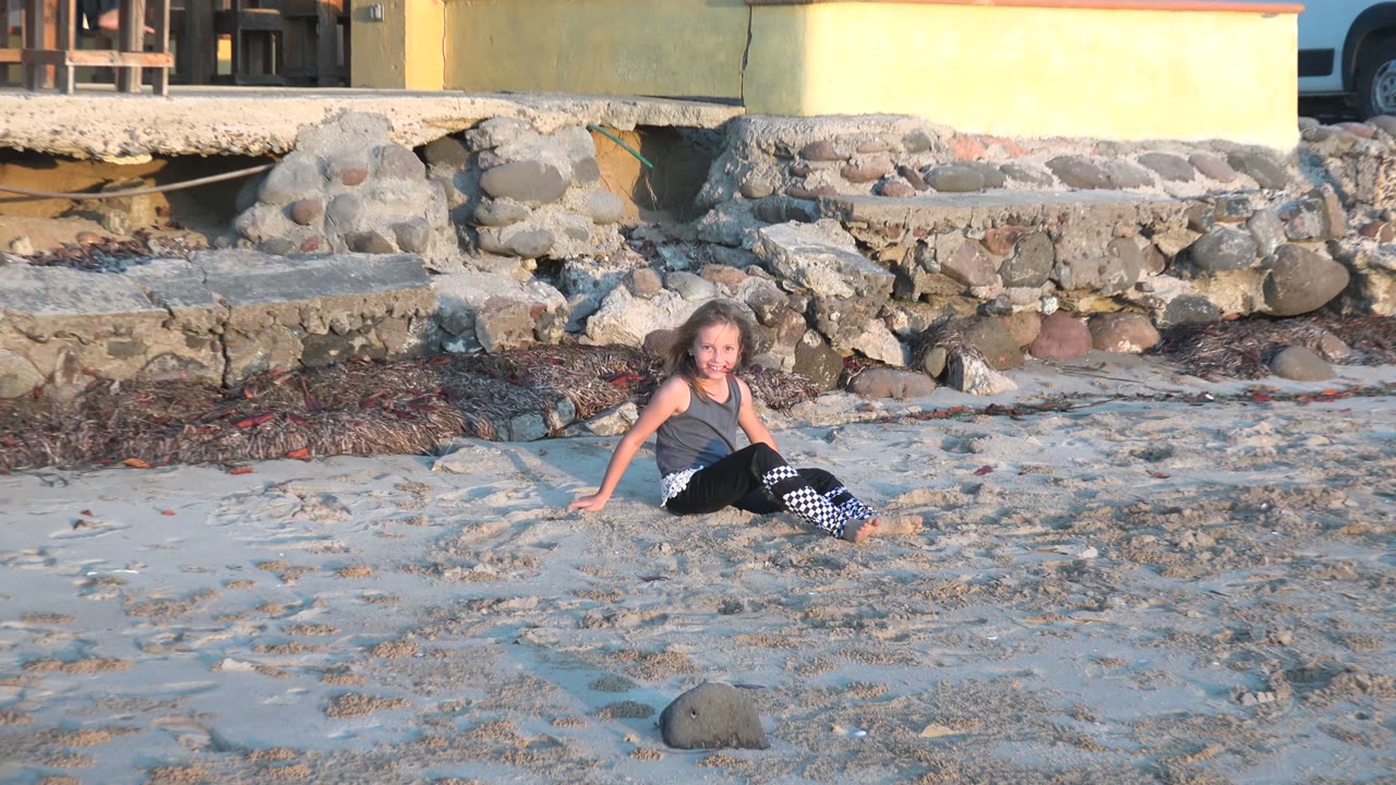 At 0:02, the child sits on the sunlit sand in San Carlos BCS Mexico with one hand behind her, checkered pant legs visible, and rocks and seaweed behind.