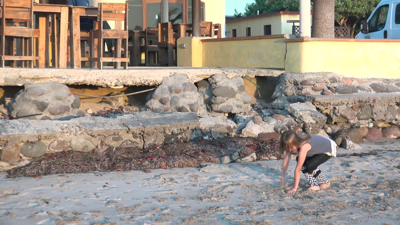 At 0:00 in San Carlos BCS Mexico, a young girl crouches on the sandy beach while adults stand near outdoor seating above a stone wall in warm light.