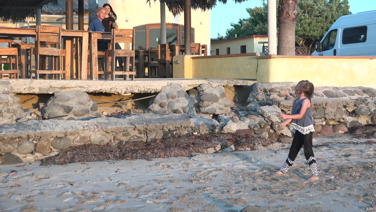 Girl walking on sandy beach with stone wall, van, and people behind her.