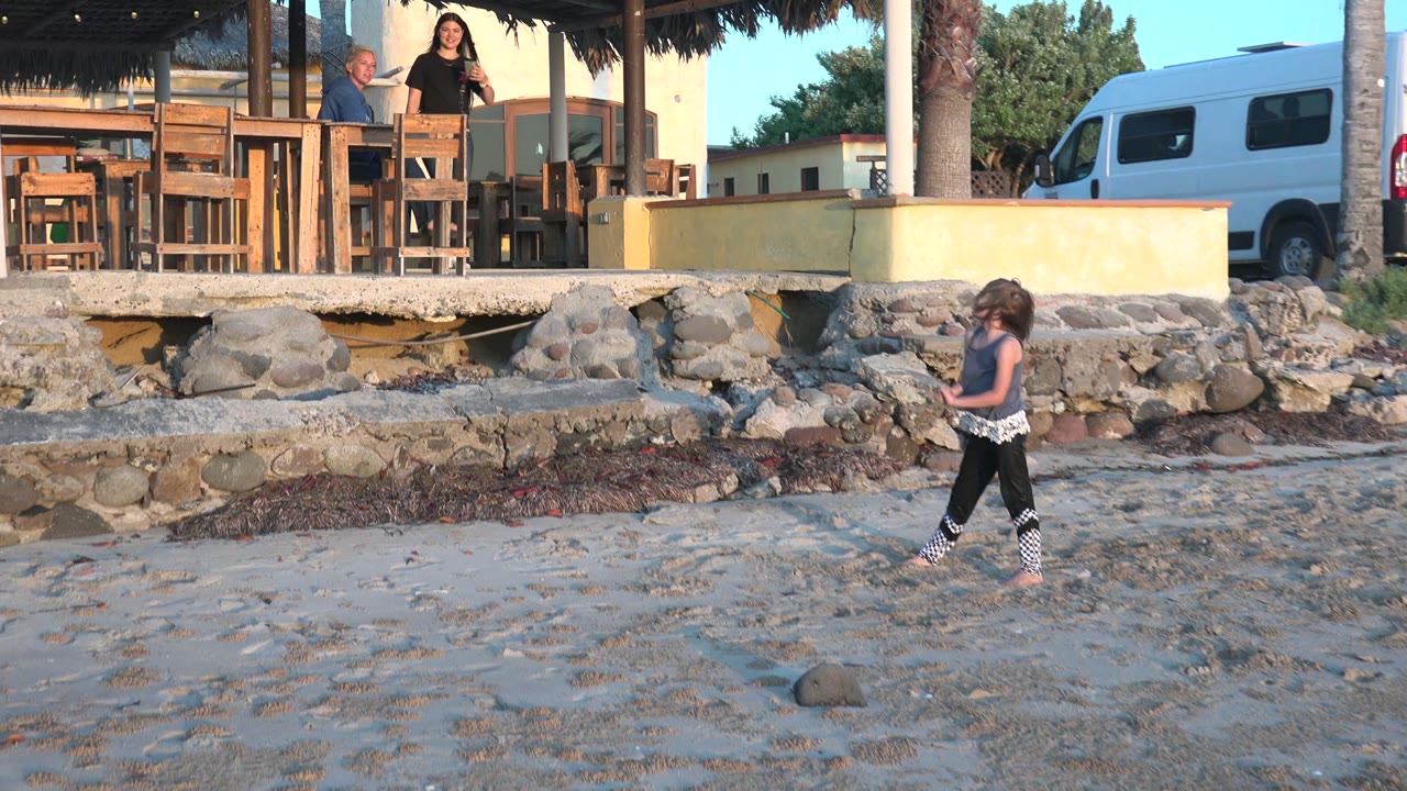 At 0:02 in San Carlos BCS Mexico, the girl walks farther right across rippled sand while adults stand by the raised outdoor seating above the stone wall.