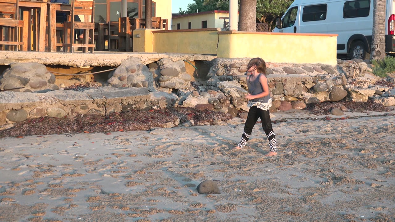 A girl in a dark top and black pants moves across sandy beach in San Carlos BCS Mexico, with rocks, a yellow wall, outdoor furniture, and a parked van behind her.
