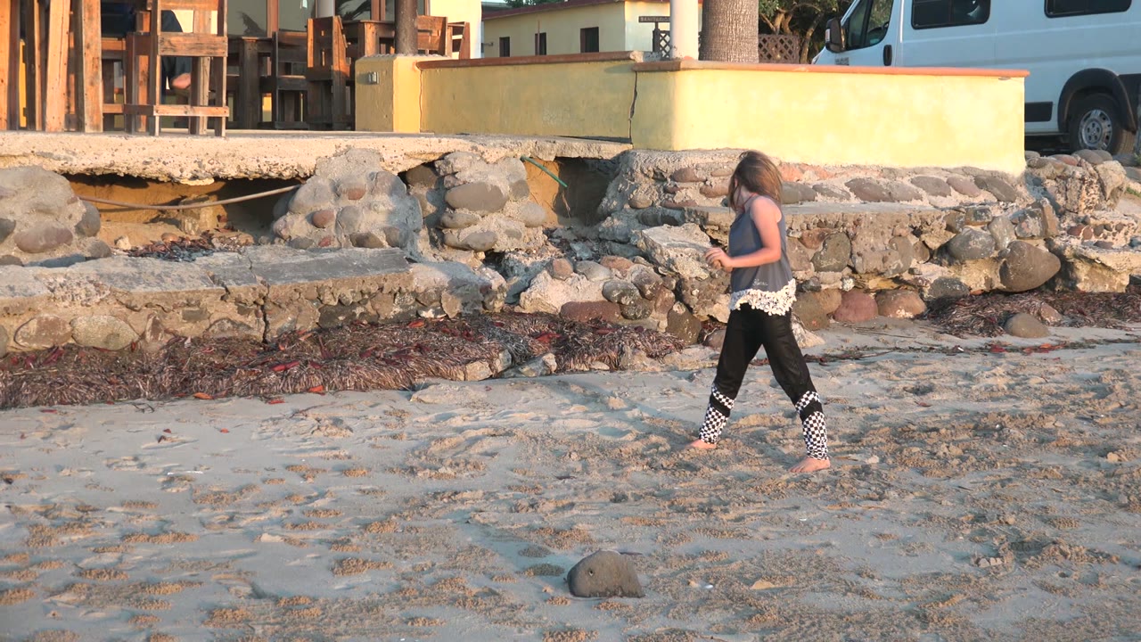 The girl pauses mid-step on the sandy San Carlos BCS Mexico beach, framed against rocky shoreline work, a yellow wall, and a parked van near a building.